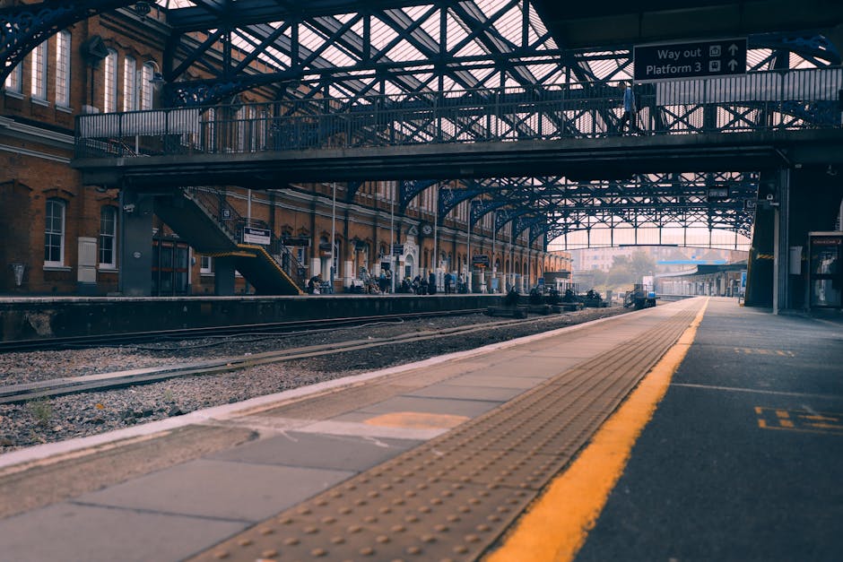 A commercial cleaning worker dressed in a high-visibility yellow vest with reflective stripes is walking down a staircase at Sutton station, carrying a green cleaning bucket and a mop. The staircase h